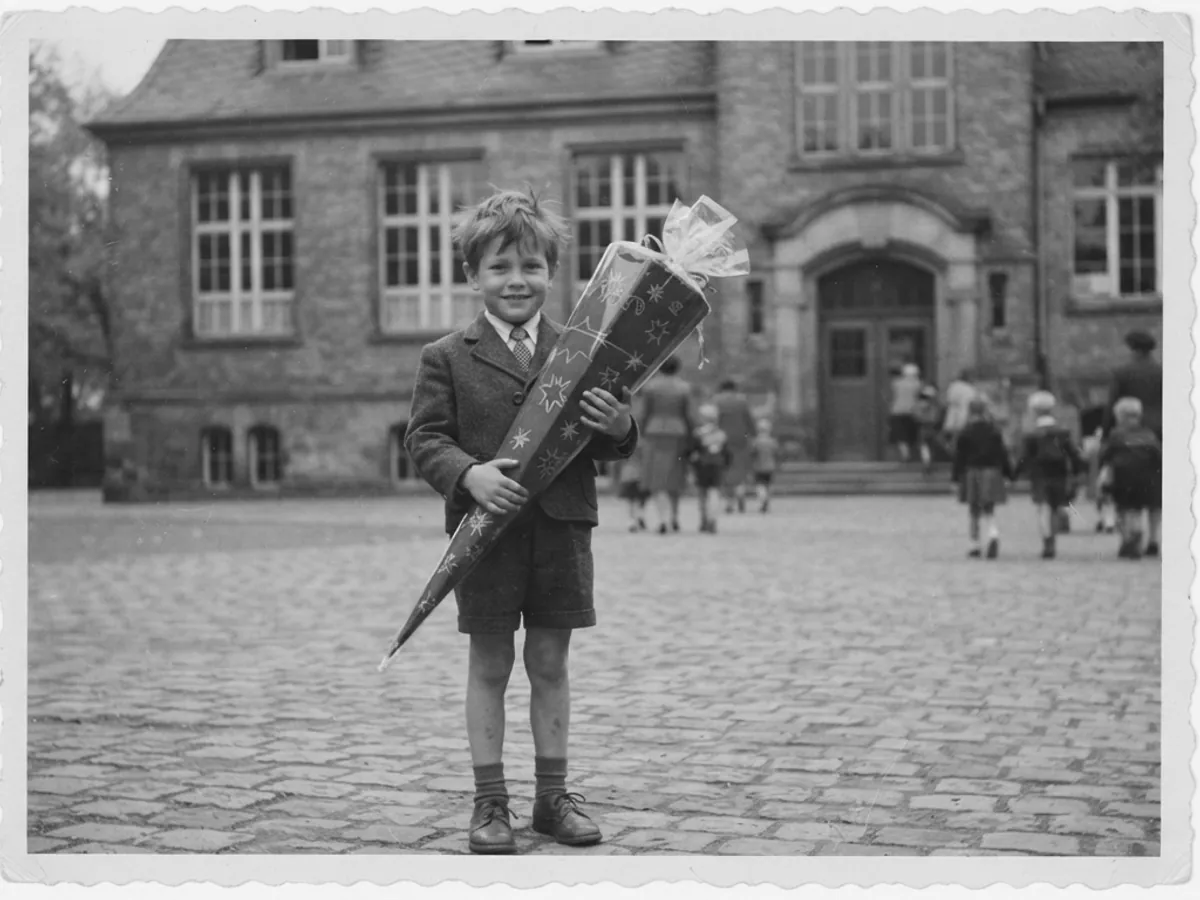 A black-and-white photo of a boy with a school cone in front of the school